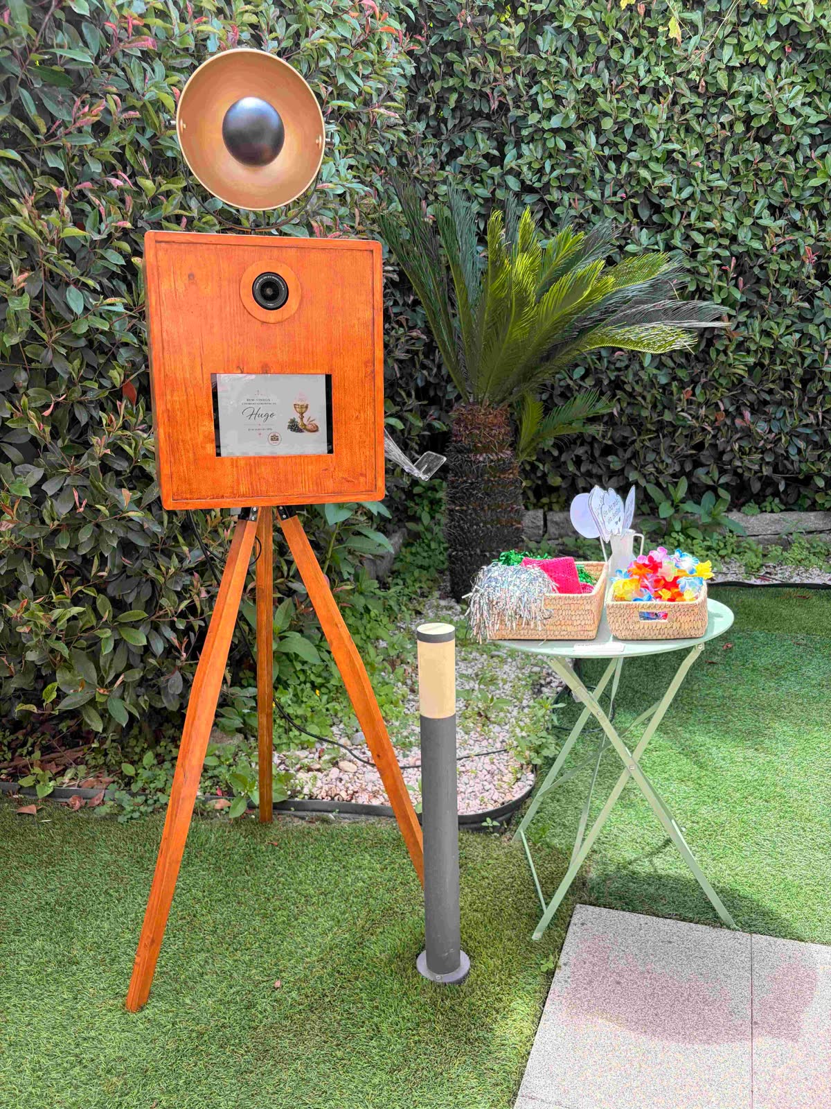 Photobooth montado num jardim sobre relva, com uma mesa de apoio e um caixote de adereços coloridos ao lado