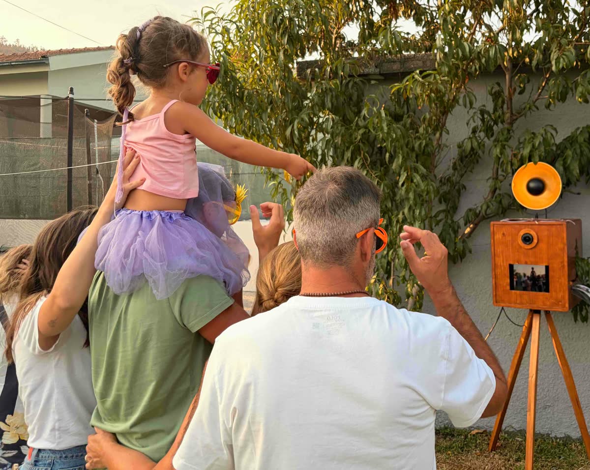 Família a posar com adereços junto ao photobooth num jardim, com uma criança de tutu e óculos em forma de coração ao colo