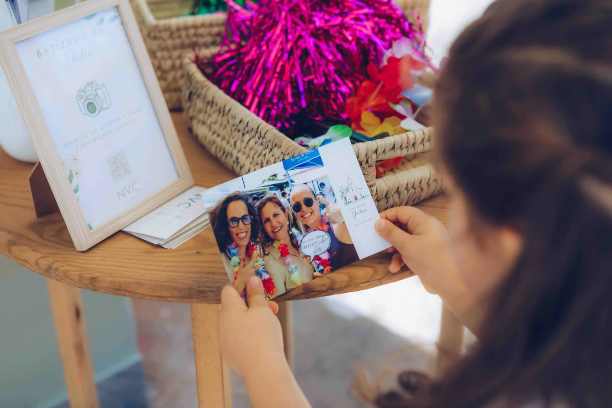 Convidada de um batizado a segurar a fotografia acabada de imprimir, com o template personalizado do evento, ao lado do cesto de adereços com colares havaianos e pompons